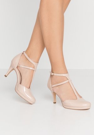 Jambes portant des talons aiguilles en cuir verni beige avec bride en T et fines boucles à la cheville sur une surface blanche.