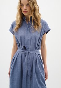 Rochie cămașă de culoare albastru deschis, cu nasturi pe față, mâneci scurte și talie legată. Țesătura are dungi texturate subtile.