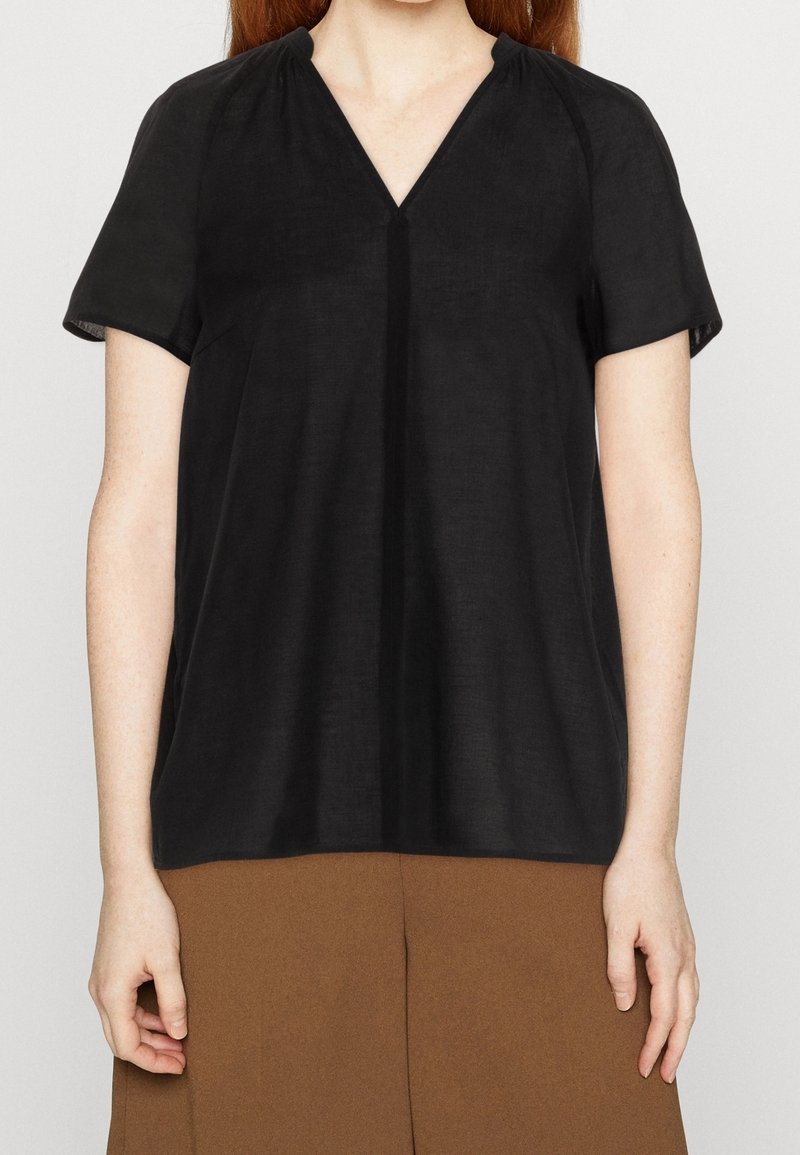 Femme portant un chemisier noir uni à manches courtes et col en V, avec un pantalon marron, debout devant un fond clair uni.