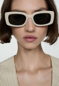 Beige rectangular sunglasses with dark lenses, featuring a thick frame. The wearer has a neutral expression and short brown hair. Striped top visible.