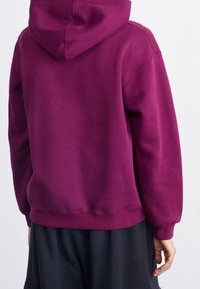 Sudadera púrpura hecha de tejido suave, con una gran capucha, puños acanalados y dobladillo. La parte trasera es lisa con mínimos detalles de costura visibles.