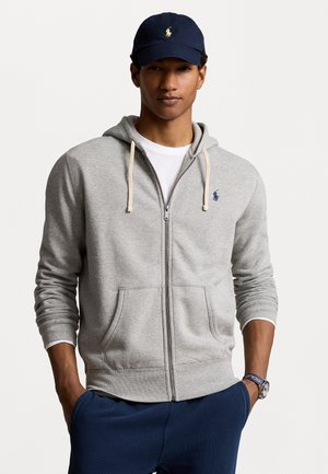 Felpa grigia con zip, cordini color crema, tasche frontali e piccolo logo blu. Abbinata a pantaloni blu navy e un cappellino blu navy con logo giallo.