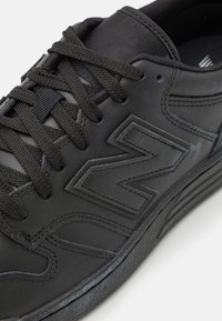 Baskets noires New Balance avec lacets texturés, logo cousu sur le côté et dessus en cuir lisse, montrées en gros plan.