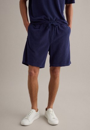 Uomo che indossa pantaloncini con coulisse blu navy e camicia abbinata, in piedi con le mani nelle tasche, abbinati a sneaker bianche su sfondo neutro.