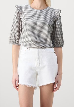Kvinde iført en sort- og hvidternet bluse med flæser på skuldrene og hvide denimshorts med frynset kant.