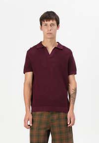 Polo en tricot bordeaux avec un col en V et des manches courtes, associé à un pantalon à carreaux marron avec des accents verts et une coupe décontractée.