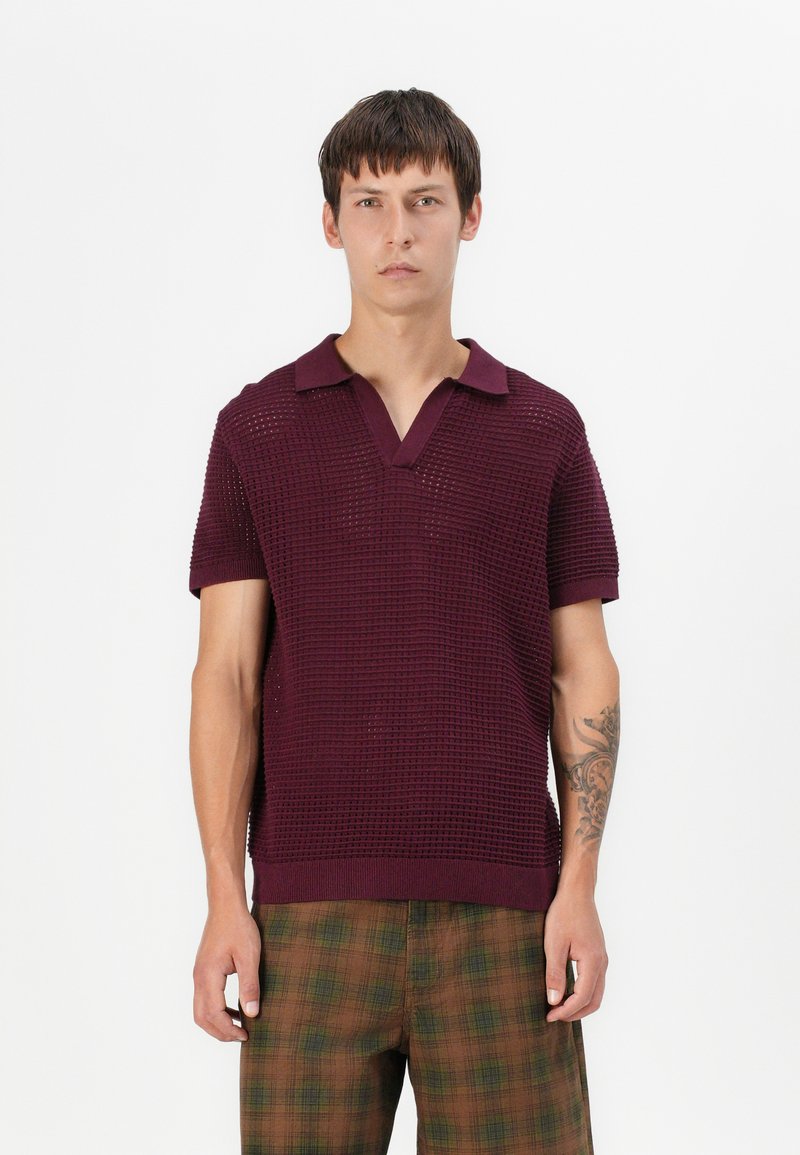 Polo en tricot bordeaux avec un col en V et des manches courtes, associé à un pantalon à carreaux marron avec des accents verts et une coupe décontractée.