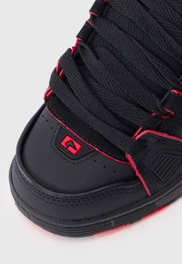 Black sneaker featuring red accents, textured laces, and a curved toe cap. Signature logo in red on the upper side. Rubber outsole with tread.