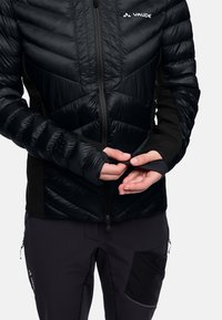 Ingrandimento di una persona che regola la manica di una giacca Vaude imbottita nera con foro per il pollice, indossando pantaloni outdoor neri con tasca con zip.