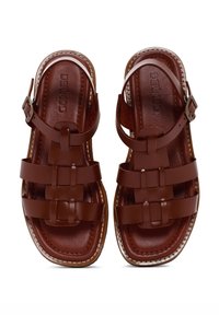 Sandales en cuir marron avec des brides tressées, une boucle réglable à la cheville et une semelle intérieure texturée avec un surfil blanc contrastant.