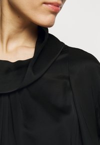 Gros plan sur le bas du visage et le cou d'une personne portant un vêtement noir avec un large col plié, sur un fond clair uni.