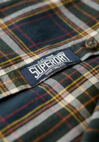 Tissu à carreaux en tons bleu marine, vert et jaune avec une étiquette "Superdry" détaillée cousue sur un fond bleu marine contrastant. Bouton marron visible.