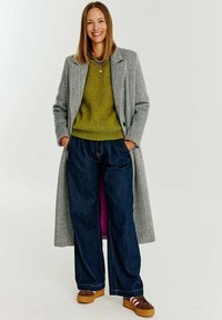 Dlouhý kabát v houndstooth vzoru, olivový svetr, volné tmavé džínsy a hnědé tenisky s bílými pruhy. Minimalistické šperky jako akcenty.