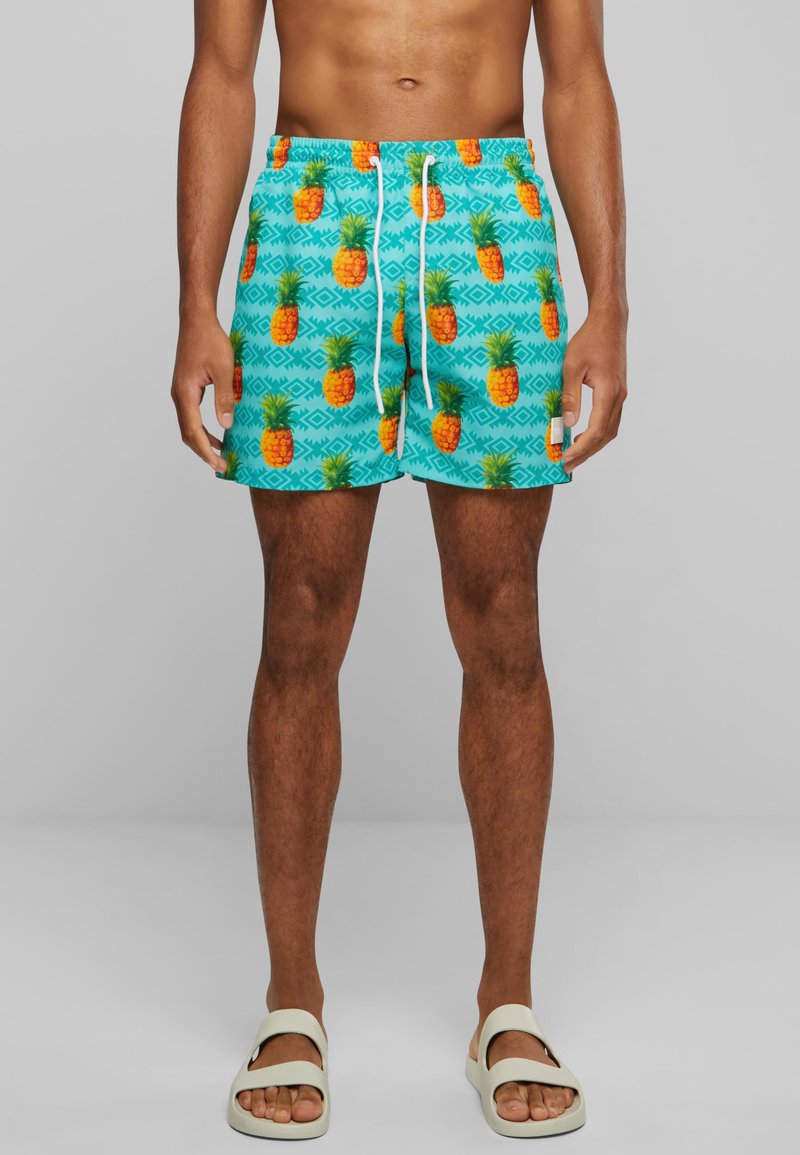 Shorts de bain turquoise avec un motif d'ananas, taille élastique avec cordon de serrage et coupe décontractée ; portés avec des sandales de couleur claire.