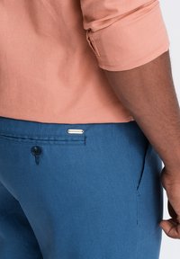 Une chemise orange clair à manches retroussées à côté d'un pantalon bleu avec une poche avant et un bouton, arborant une étiquette dorée portant la mention "QUINTESSENCE".