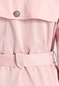 Jasnoróżowa kurtka z dużym kołnierzem, zapięciem na guziki i paskiem zawiązywanym w talii. Tkanina ma gładką fakturę.