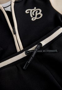Survêtement noir et beige avec zip et cordon, comprenant un logo brodé "TB" et la mention "Baker by Ted" imprimée sur le cordon.