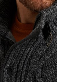 Suéter de punto gris oscuro con textura de canalé, que presenta un cuello alto, ribetes negros y un acento de metal en el cuello.