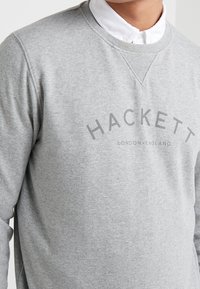 Sudadera gris con mangas largas, cuello redondo y acabado texturizado. Presenta "HACKETT LONDON-INGLATERRA" impreso en gris oscuro.