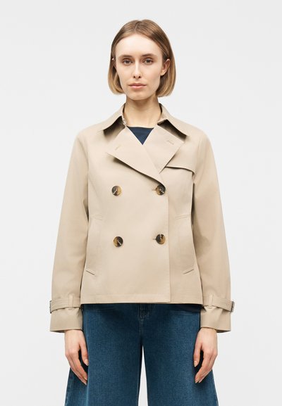Femme portant un trench-coat beige croisé à larges revers et un jean bleu, debout devant un fond clair uni.