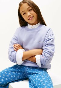 Lilla sweatshirt med ribbede mansjetter og et logo på brystet, parret med lyseblå bukser med blomsterprint og en hvid skjorte under.