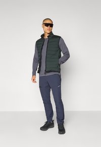 Gilet imbottito verde scuro sopra una maglietta a maniche lunghe grigia, abbinato a pantaloni blu notte e scarpe da trekking nere. Presenta tasche con zip e un design elegante.