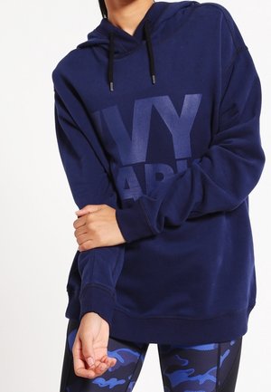 Osoba nosząca granatową bluzę z kapturem z dużym napisem "IVY PARK" oraz niebieskie legginsy w kamuflażowy wzór, poprawiająca rękaw bluzy z założonymi rękami.