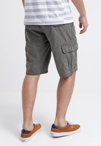 Pantaloni cargo verde oliva con molte tasche, lunghezza a metà coscia e vestibilità rilassata. Indossati con scarpe da ginnastica beige con dettagli blu.