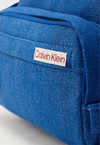 Μπλε τζιν σακίδιο με λεία υφή, με μια ετικέτα Calvin Klein σε κόκκινο και λευκό ραμμένη στο πλάι.