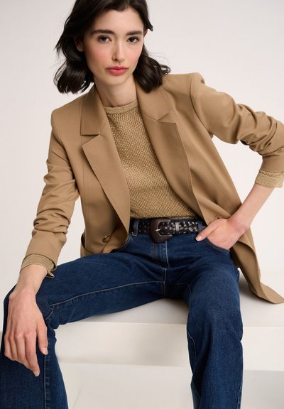 Tan blazer over a textured gold top, paired with dark blue jeans. A woven black belt accentuates the waist, highlighting the outfit's design.
