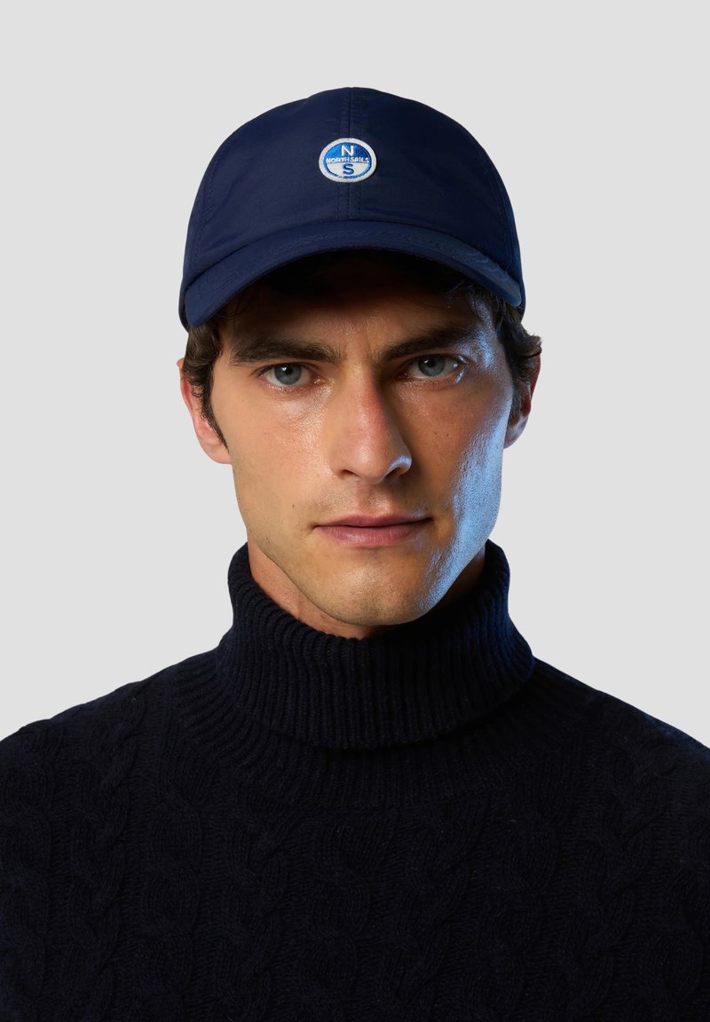 Cappellino da baseball navy con logo rotondo ricamato sulla parte anteriore, caratterizzato da un design strutturato e un tessuto leggero. Indossato con un dolcevita lavorato a maglia scuro.