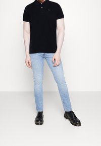 Schwarzes Poloshirt mit Knopfleiste, hellblaue schlanke Jeans und schwarze Schnürstiefel mit gelben Akzenten. Einfarbiger Hintergrund.