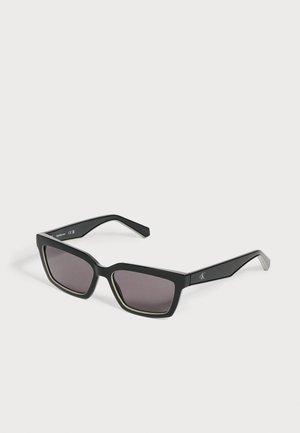 Μαύρα ορθογώνια γυαλιά ηλίου με σκουρόχρωμους φακούς και διακριτικό λογότυπο στο κλασικό, εμφανιζόμενα σε απλό ανοιχτόφωτο φόντο.