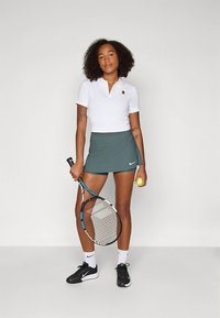 Vit pikétröja med krage, kombinerad med en grön tennis kjol. Håller en tennisracket och boll, bär svarta sneakers och vita strumpor.