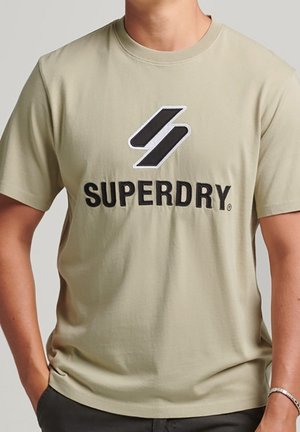 Homme portant un t-shirt beige Superdry avec un logo noir et blanc centré sur la poitrine, associé à un pantalon foncé et un bracelet argenté.