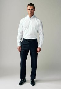 Camisa blanca de botones combinada con pantalones oscuros de corte ajustado, cinturón de cuero negro y zapatos de vestir negros brillantes. Diseño simple y clásico con detalles mínimos.