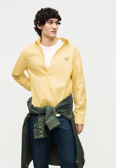 Νέος άνδρας με σγουρά σκουρόχρωμα μαλλιά, που φοράει κίτρινο πουκάμισο με κουμπιά πάνω από λευκό μπλουζάκι, μπλε τζιν και τζιν μπουφάν δεμένο γύρω από τη μέση του.