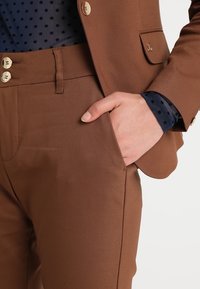 Traje marrón a medida con botones metálicos. Vista en primer plano del bolsillo del pantalón y la mano, superpuesto con una blusa de lunares azul marino por debajo.