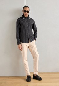 Gestreifte schwarze Hemdbluse mit Kragenakzenten, hellbeige Hosen und schwarzen Schuhen, die einen modernen Schnitt und eine entspannte Passform zeigen.