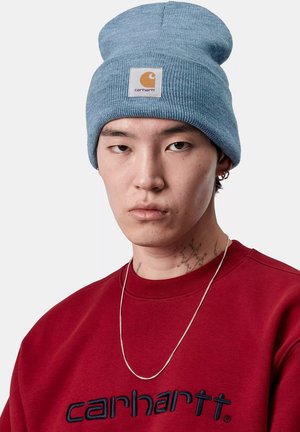 Niebieska dzianinowa czapka z daszkiem z naszywką z logo Carhartt; noszona z czerwoną bluzą z niebieskim haftowanym napisem i srebrnym naszyjnikiem.