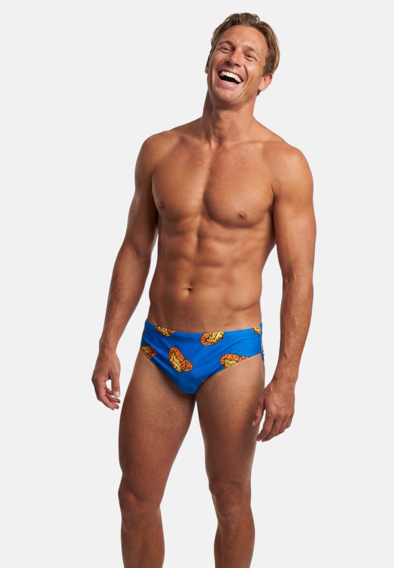 Hymyilevä lihaksikas mies, joka pukeutunut sinisiin uimahousuihin, joissa on oransseja sushirullaprinttejä, seisoo valkoista taustaa vasten.