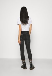 Mujer con cabello largo y oscuro de pie de espaldas, vestida con una camiseta blanca, jeans negros con bordados coloridos en los puños y botas negras.