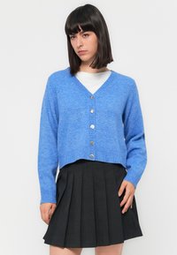 Blauer Wollmischung-Kardigan mit V-Ausschnitt, langen Ärmeln, silbernen Knöpfen und gerippten Bündchen, kombiniert mit einem gefältelten schwarzen Rock.