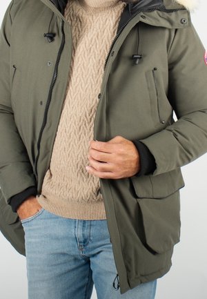 Homme portant un jean bleu clair, un pull beige en maille torsadée et une veste d’hiver vert olive avec une capuche bordée de fourrure, ajustant la fermeture éclair de la veste.