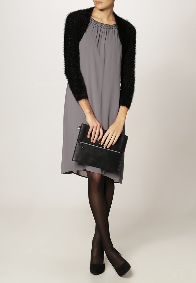 Femme en robe grise, boléro noir en matière duveteuse, collants noirs et talons, tenant une pochette noire devant un fond neutre.
