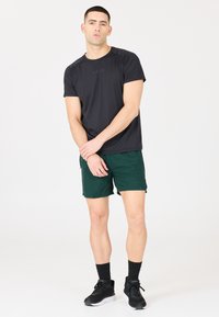 Musta urheilupaita lyhyillä hihoilla, yhdistettynä tummiin vihreisiin shortseihin. Kevyt kangas, rento istuvuus ja mustat lenkkarit valkoisella pohjalla.