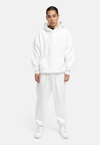 Weißer übergroßer Kapuzenpullover mit einer Kängurutasche, kombiniert mit passenden Jogginghosen. Glattes Material, lässige Passform und Kordelzugdetails.