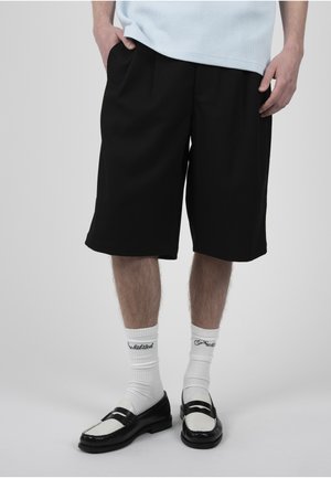 Homme portant un short large noir, des chaussettes blanches avec du texte et des mocassins noir et blanc, debout les mains dans les poches.