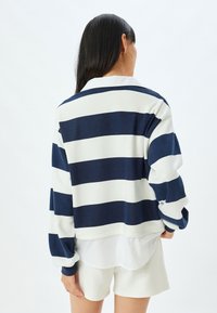 Maglione a righe blu navy e bianche con maniche lunghe, vestibilità comoda, tessuto testurizzato e scollo rotondo. Indossato sopra una camicia bianca.