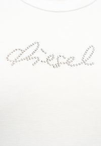 Ασημένιες στρας διατεταγμένες ώστε να σχηματίζουν τη λέξη "Ariesel" με καλλιγραφικά γράμματα σε φόντο λευκού υφάσματος.